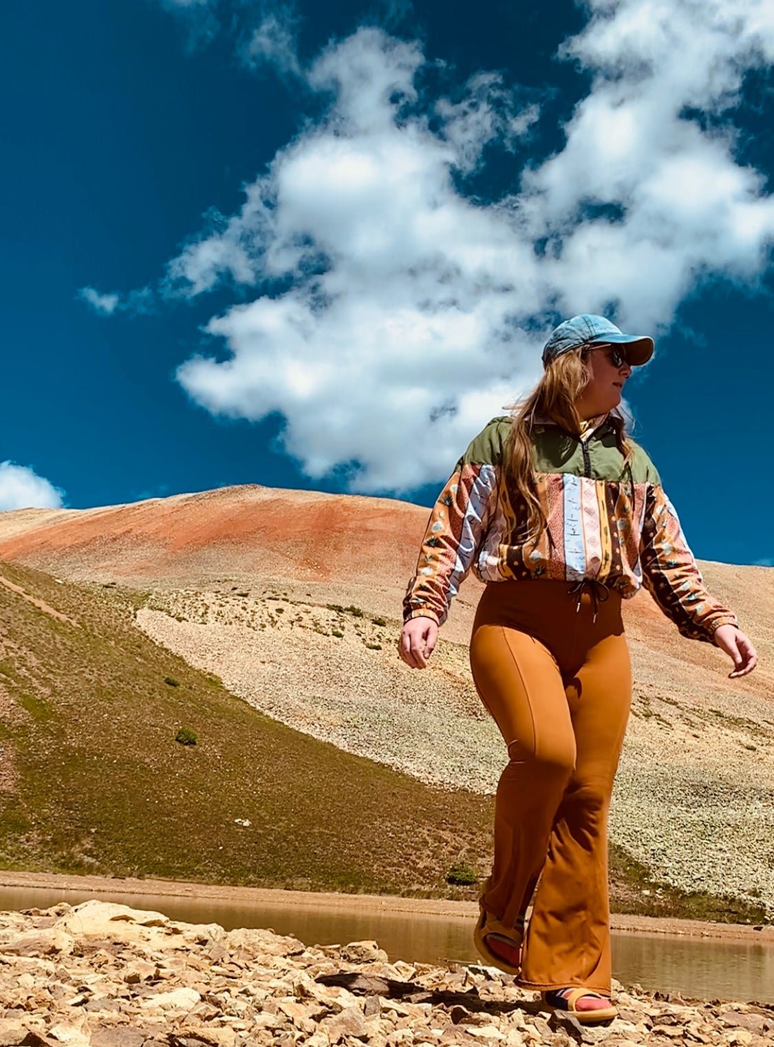 Ali Harris walking near a small pond at the top of an off-road trail in Ouray, Colorado. A colorful mountain peak and a blue sky make for a gorgeous backdrop.