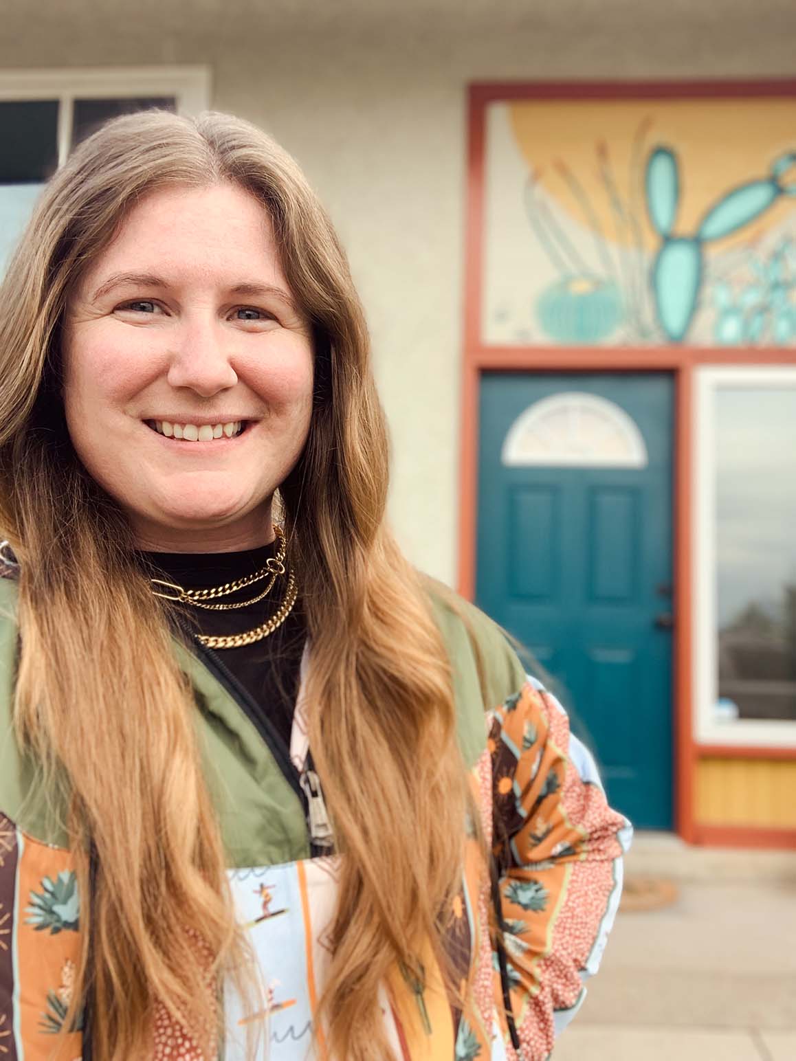 Ali Harris in her Desert Boarder Print Windbreaker. In the background you can see the mural she painted above her front porch, which is a desert plant scene featuring various types of cacti, an ocotillo, and a banana yucca.