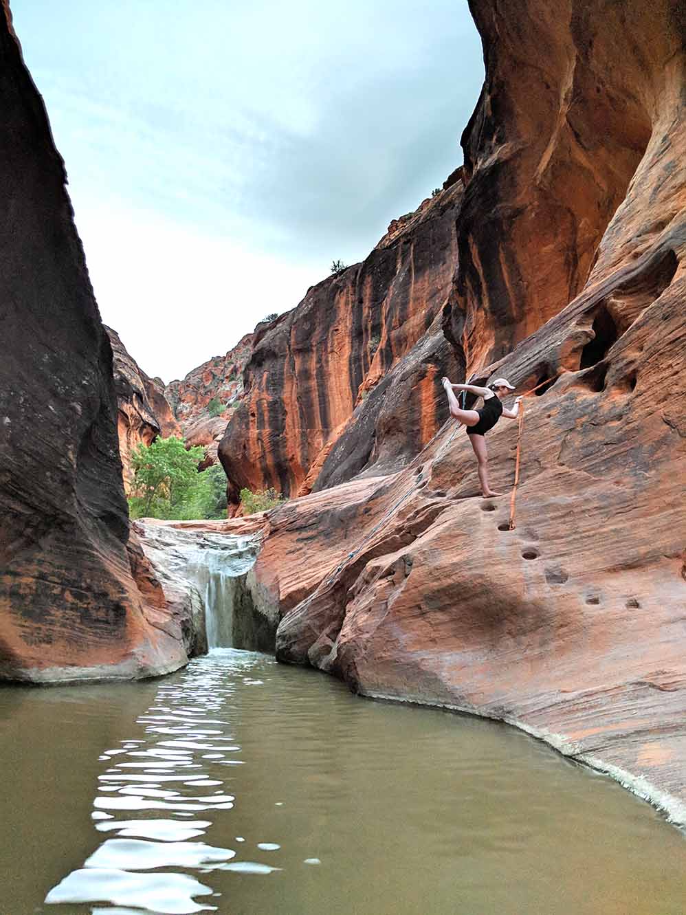 Ali is holding on to the rope attached to the cliffside for this hike to assist hikers up the rock to the top of a small waterfall. Ali is doing dancer's pose. In the background you can see the red and black rock cliff faces of the canyon the waterfall is coming out of. In the foreground, is the small pool created by the waterfall.