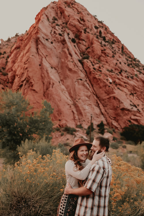 Ali and her husband are embracing and laughing. Behind them, you can see rabbit brush in full bloom, and a red rock mountain peak.