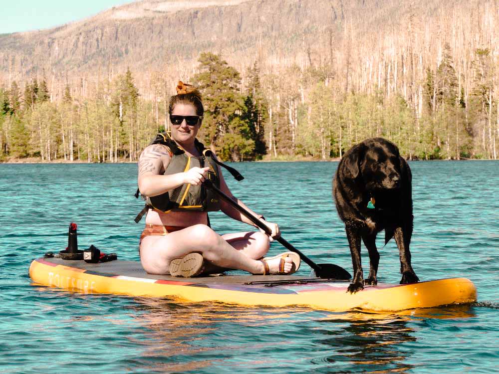 Ali Is sitting on her paddle board paddling, while her black lab collie mix dog, Meru, is standing at the front of the board. Both Ali and Meru are facing the camera. Ali is wearing her PFD, with good reason. Shortly after this photo was taken, Meru lept off the board to shore, dumping Ali in the water.