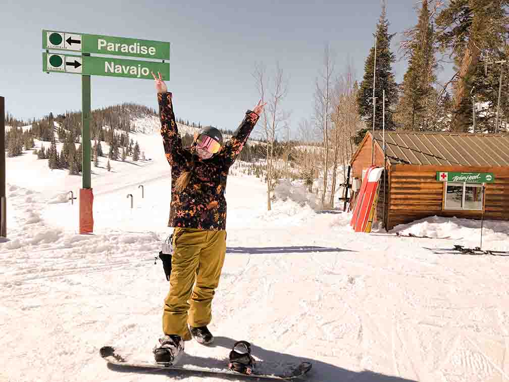 Ali is standing at the top of the the slope with one boot clipped in her snowboard and one boot out. She is holding up both of her arms making peace signs with her hands. Her coat is a black, coral, tan, gold, and orange floral print. Her snow pants are a gold color. She has her helmet and googles on. Behind her, you can see a small mountain peak (because she is already close to the top of the mountain), and sign pointing towards the Navajo ski run, and a small log cabin used as a first aid station.